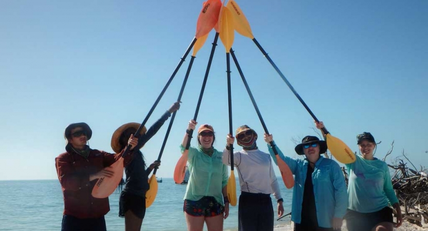 A group of people touch lifted paddles together on a beach. 